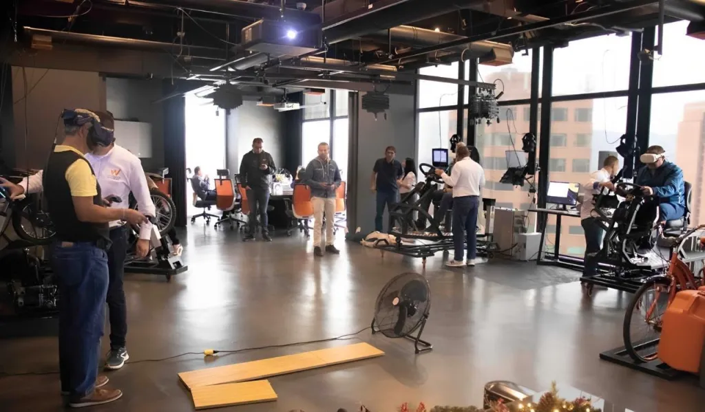 Sala de entrenamiento inmersivo con varios simuladores en uso: participantes con visor de realidad virtual practican en simuladores de moto y de conducción mientras otros asistentes observan.