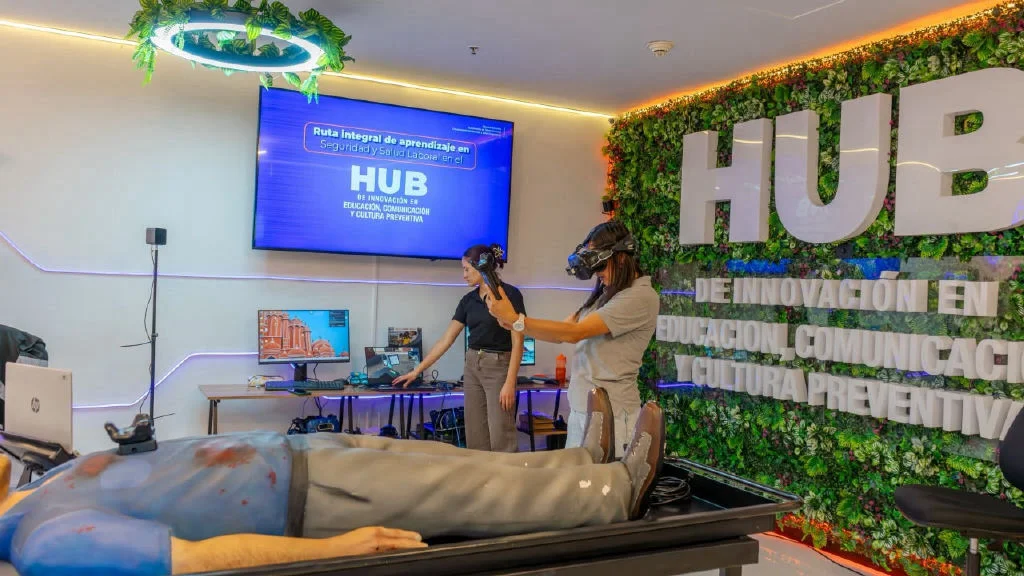 Sala HUB de WayGroup: una participante con visor de realidad virtual practica junto a una instructora y un maniquí de entrenamiento en salud.